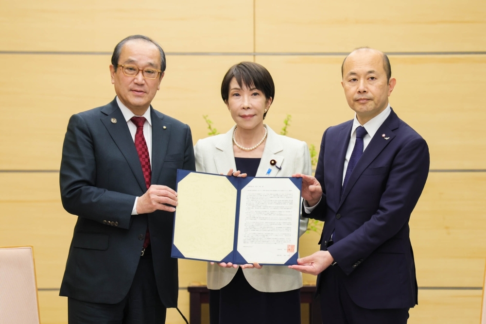 A commemorative photo of Prime Minister TAKAICHI holding up the petition and facing the camera, flanked by Hiroshima Mayor Matsui and Nagasaki Mayor Suzuki