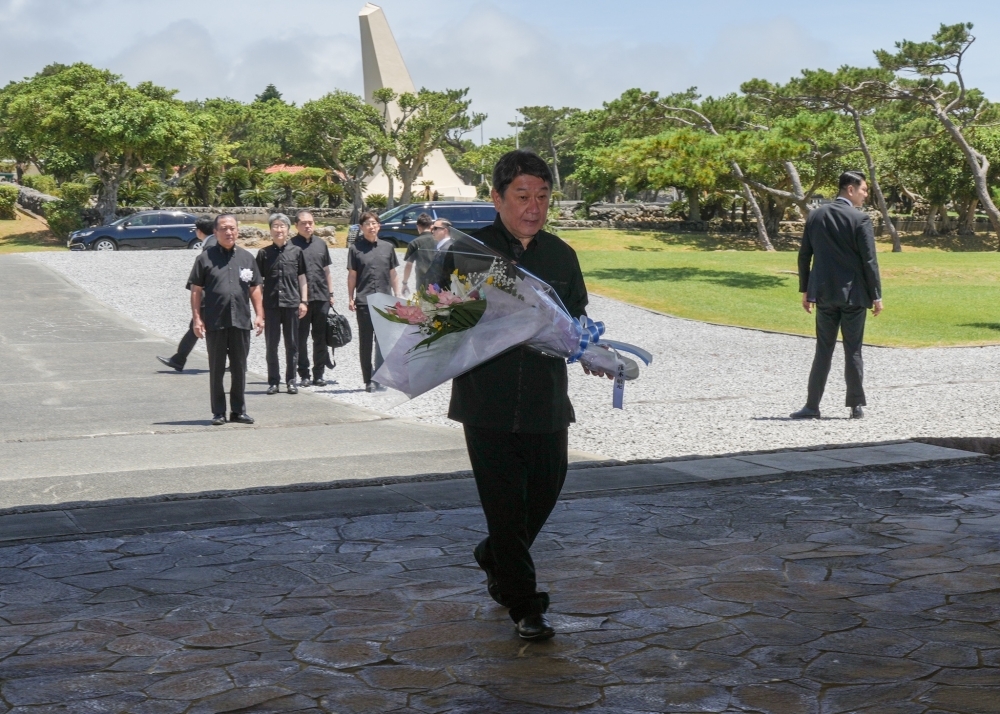 Foreign Minister Motegi, dressed in black formal attire, walking along the stone-paved path in Peace Memorial Park toward the memorial monument, carrying a large bouquet of flowers in both hands