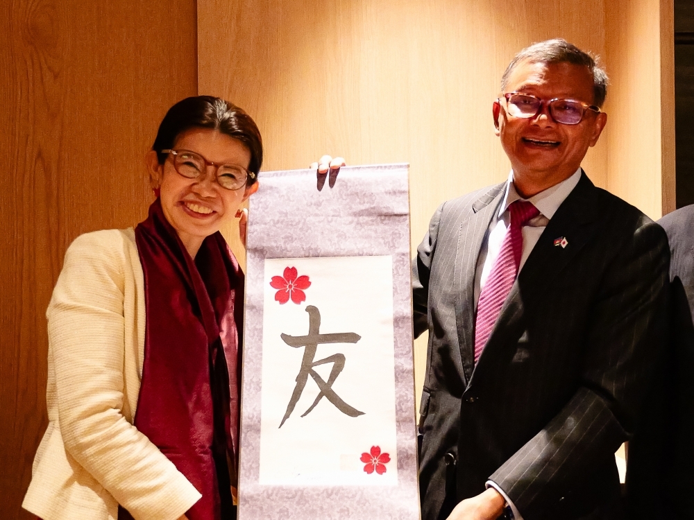 State Minister KUNIMITSU and H.E. Dr. HANG, Deputy Prime Minister of Cambodia, pose for a photo with a hanging scroll inscribed with the word 'Friend' between them