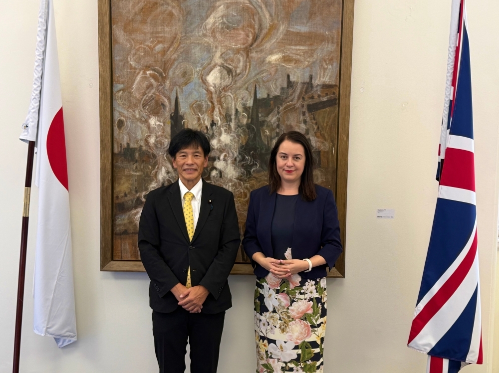 Parliamentary Vice-Minister SHIMADA taking a picture with Ms. Stephanie Peacock MP, Parliamentary Under-Secretary of State, Department for Digital, Culture, Media and Sport, in front of  a landscape picture and national flags