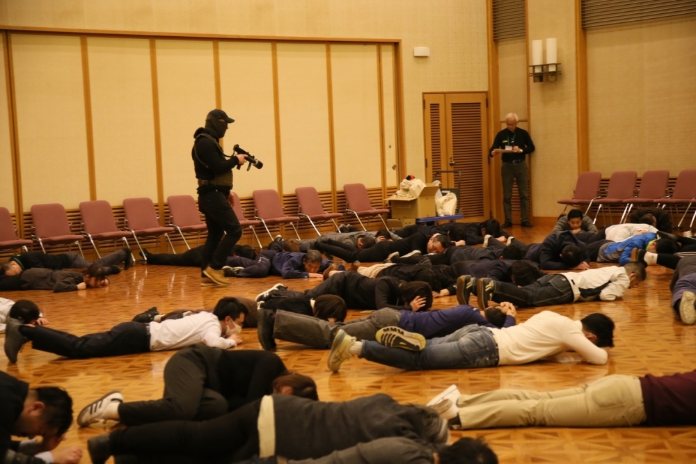 銃を持った犯人の命令で参加者が床に伏せる訓練の様子