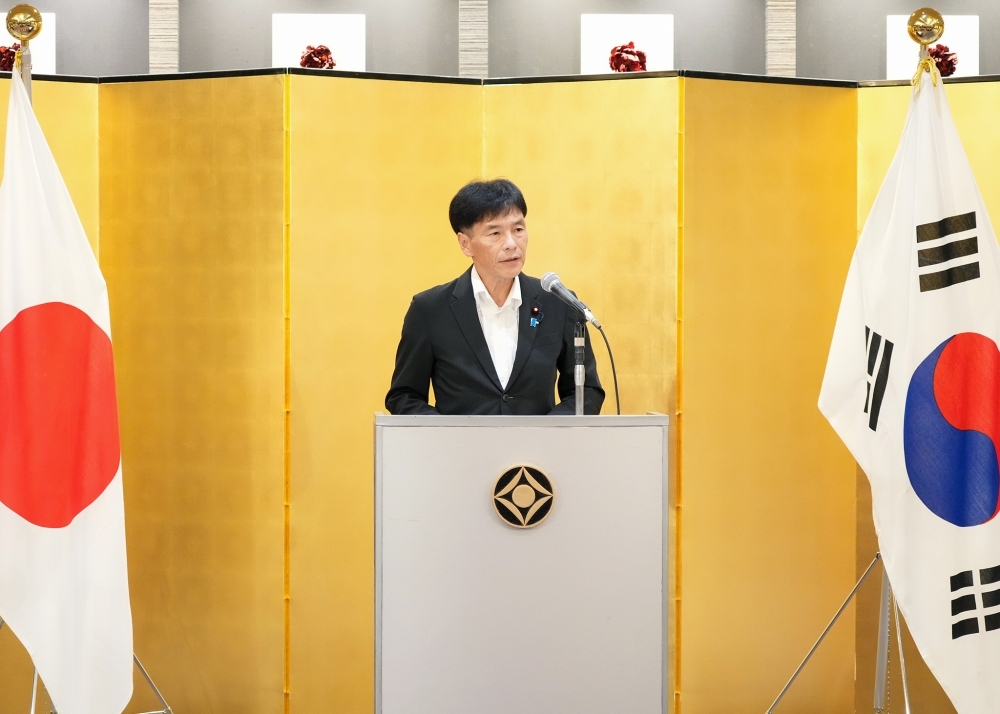 日韓両国国旗を両脇に、壇上でスピーチする島田政務官。