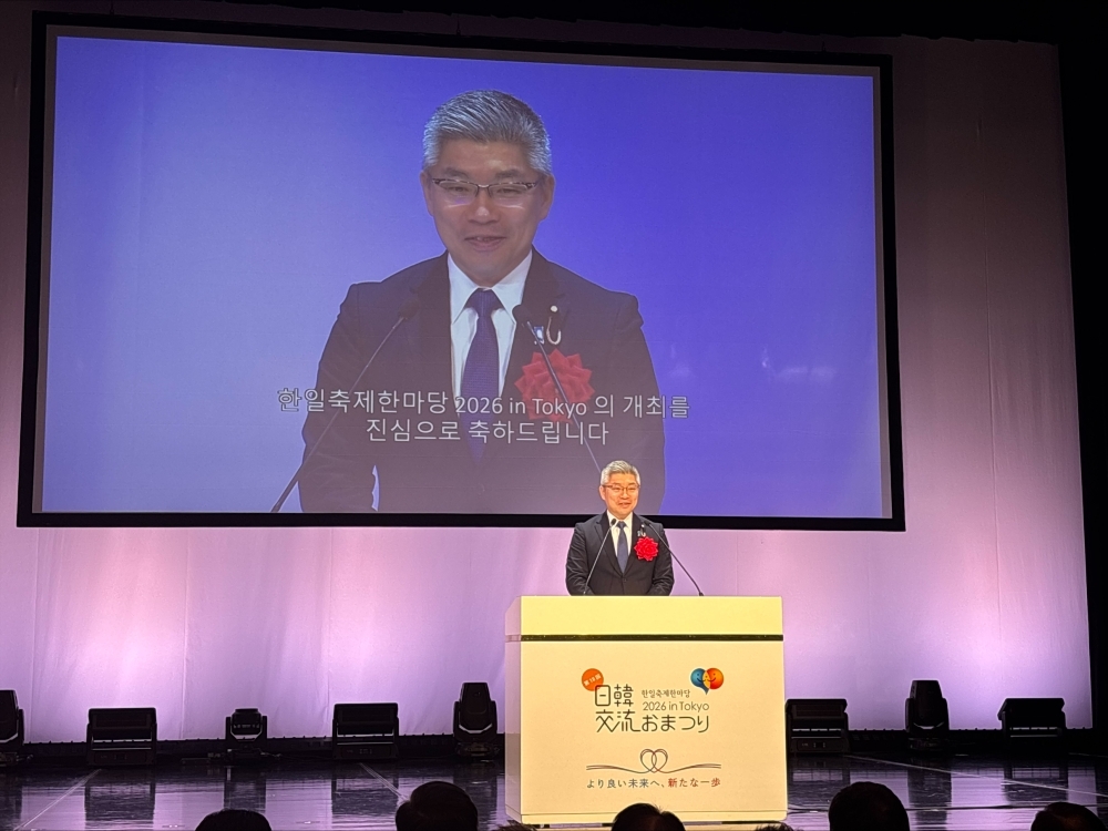 日韓交流おまつりの開会式において、壇上で挨拶する藤井政務官。背後のモニターにも政務官が投影されている。