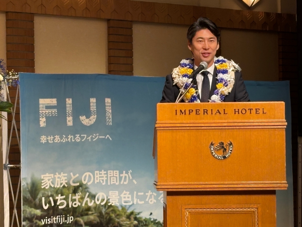 フィジーのポスターを背後に、花の首飾りをつけて壇上でスピーチする中曽根副大臣。