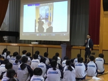 宮本講師が会場前方のスクリーンの横で立って生徒に説明する様子。