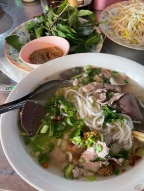 手前は器に鶏ガラスープの餅米麺が入った様子。奥に付け合わせの葉っぱ類やもやしなど。