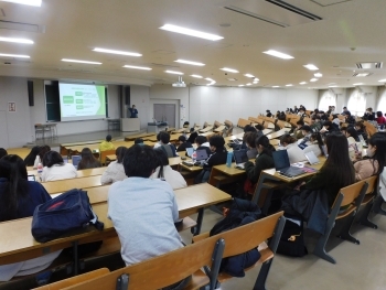 教室内で、大学生がスクリーンに投影された資料を見ながら、講師の説明に耳を傾けている様子。