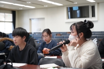 教室内で学生がマイクを持ち、講師の問いかけに対して意見を述べている様子。