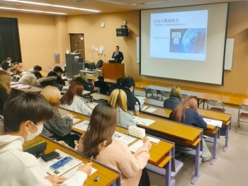 教室後方から前方を写した様子。学生が講師の説明を聞きながら資料に目を通し、メモを取りつつ講義に集中する場面。