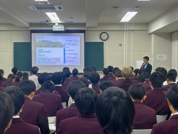 教室前方で講師がスライドを用いながら、生徒に向けて説明を行い、教室には多くの生徒が着席し、講義を聞いている。