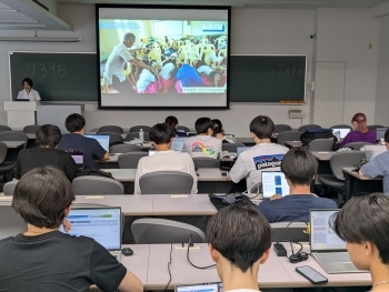 教室で学生がノートパソコンを開き、オンラインで外務省ODA出前講座を受講する様子。前方のスクリーンには講座映像が映し出されている。